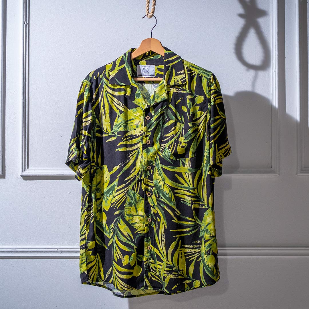 A short-sleeved shirt with a tropical leaf print hanging on a hanger against a dark background.